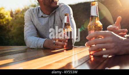Giovane seduti all'aperto al tavolo di legno avente le birre e parlare. Focus sul giovane mani con bottiglie di birra durante una festa all'aperto. Foto Stock