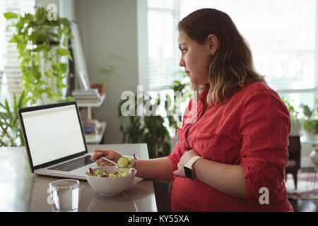 Donna incinta avente la prima colazione durante l'utilizzo di laptop Foto Stock