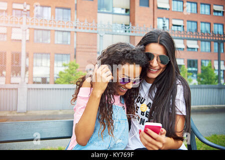 Due ridendo giovani fidanzate seduta su una panchina della città in estate per ascoltare musica insieme su un comune paio di cuffie Foto Stock
