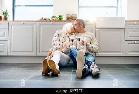 Affettuosa coppia senior kissing seduti insieme sul piano della loro cucina a casa a bere il vino rosso Foto Stock