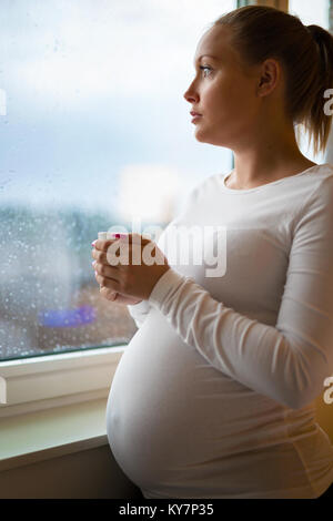 Premurosa donna incinta guardando fuori dalla finestra drinkng tea Foto Stock