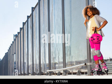 Ritratto di giovane ragazza africana in abbigliamento protettivo in piedi accanto alla recinzione di skate park Foto Stock