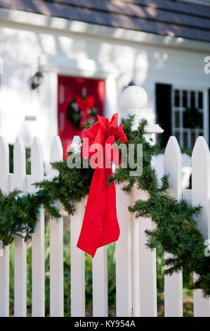 Ghirlanda con fiocco rosso appeso su bianco Picket Fence tradizionale circostante, bianco, casa coloniale, corona in background Foto Stock