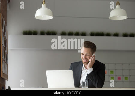 Imprenditore sorridente indossare tuta seduti alla scrivania tramite digitazione su laptop, rendendo di risposta a una chiamata, parlare al telefono client di consulenza circa Foto Stock