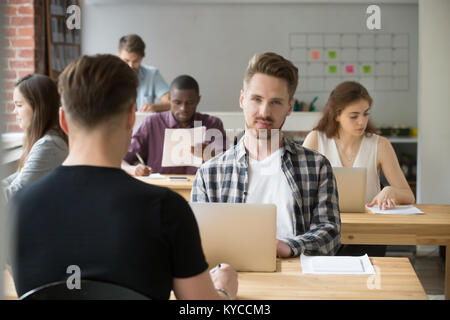 Giovane imprenditore guardando la telecamera seduto alla scrivania con computer portatile, uomo che lavora in moderno accogliente co-spazio lavoro con diverse persone e multirazziale, freelanc Foto Stock