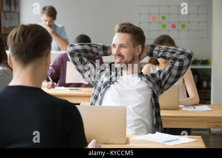 Felice giovane impreditore rilassante mani dietro la testa seduto alla scrivania con computer portatile in ufficio condiviso, uomo sorridente godendo di lavorare nel moderno e confortevole accogliente Foto Stock