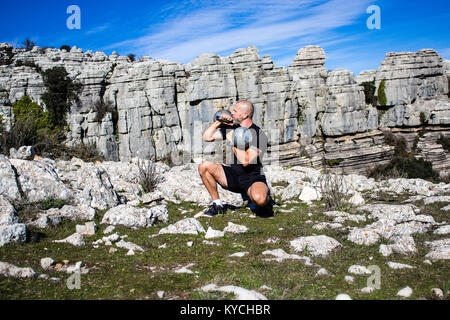 L'uomo accovacciato tenendo due Kettlebells In terreni rocciosi Foto Stock