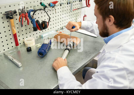 Ritratto di prosthetist al lavoro, sopra la spalla del giovane uomo Assemblaggio piede artificiale seduta a tavola nel moderno laboratorio Foto Stock