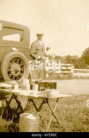 Un uomo che indossa abbigliamento formale contiene una tazza di tè e si trova dietro la sua automobile con un campo tabella impostato con diverse bocce in utensili in primo piano durante un viaggio su strada attraverso la Nuova Inghilterra, 1925. () Foto Stock