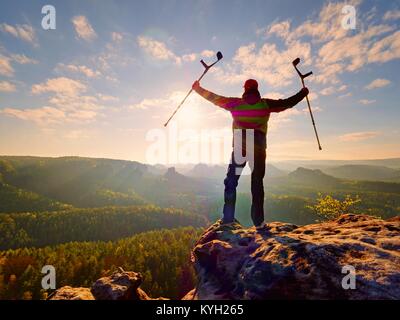 Escursionista con la medicina stampella sopra la testa raggiunto traguardo personale. Gamba rotta fissata in un immobilizzatore profonda valle misty bellow silhouette di un uomo con la mano Foto Stock