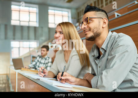 Multi-etnico gruppo di studenti seduti alla scrivania in aula magna del collegio moderno e sorridente felicemente, concentrarsi sul giovane uomo mediorientale indossando glasse Foto Stock