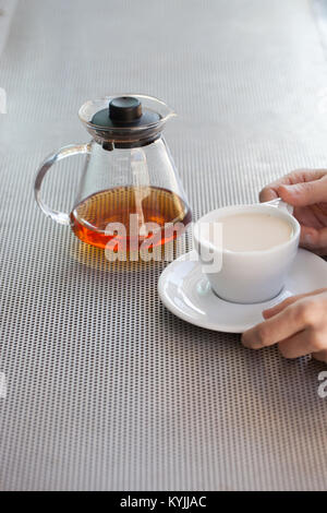 La donna caucasica mani tenere coppa in ceramica con tè nero e latte, vetro teiera in background su tavola metallica Foto Stock