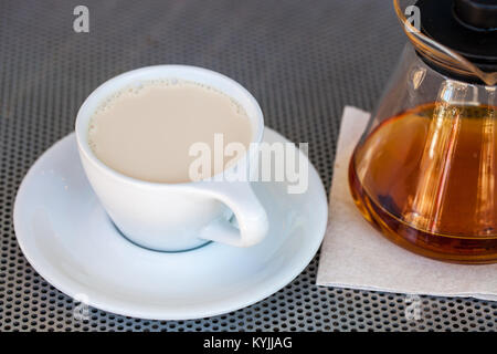 Messa a fuoco selettiva di tè nero con il latte in porcellana bianca cup con teiera accanto ad esso su una tavola metallica Foto Stock