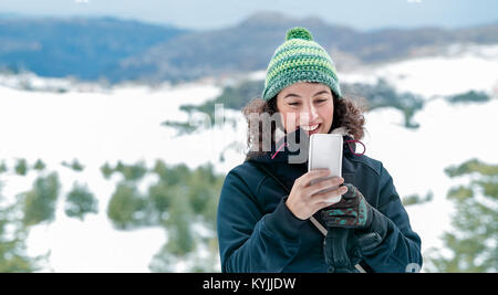 Carino donna allegra tramite telefono cellulare in montagna, ricevere messaggi divertenti, avendo divertimento all'aperto, con piacere trascorrere vacanze invernali in Foto Stock