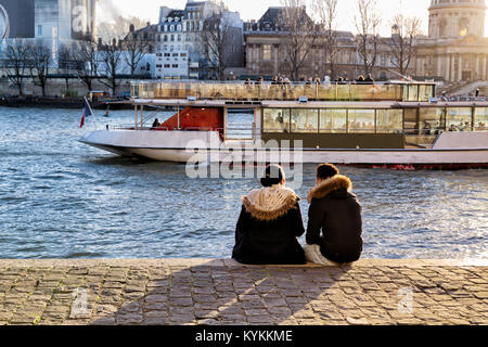 Parigi coppia giovane sedersi sul fiume Senna bank e guardare il tramonto e una barca che va da. Indossano i collari di pelliccia sul freddo giorno d'inverno. Romantica vista. Foto Stock