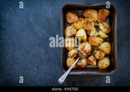 Patate arrosto con rosmarino in una teglia da forno con cucchiaio su un sfondo di ardesia. Vintage filtro applicato Foto Stock