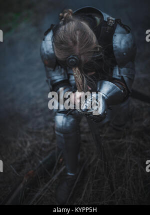 La ragazza di immagine di Jeanne d'Arc in armatura e con la spada nelle sue mani si inginocchia sullo sfondo di erba secca. Primo piano. Foto Stock