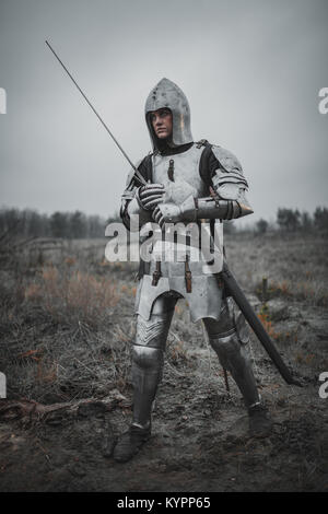 La ragazza di immagine di Jeanne d'Arc va sul prato attraverso il fango in armatura e casco con la spada nelle sue mani. Foto Stock