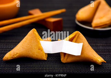 Fortune cookie con chiaro vuoto una striscia di carta, un altro biscotto e bacchette sul tovagliolo nero lo sfondo. Close-up. Foto Stock