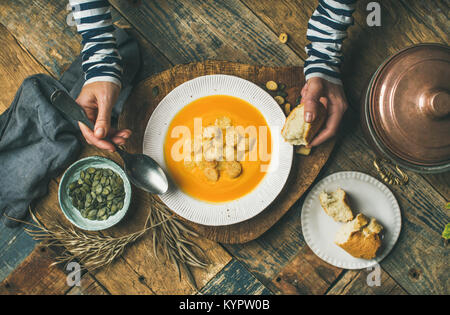 Flat-lay di mani femminili e caduta di riscaldamento Crema di zucca zuppa con crostini e semi sulla scheda sopra in legno rustico sfondo, vista dall'alto. Autunno vegetari Foto Stock