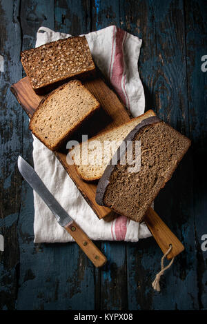 Varietà di pani fatti in casa a fette di pane di segale grano intero e semi sul tagliere di legno con carta asciugatutto e coltello sopra il vecchio scuro dello sfondo in legno Foto Stock