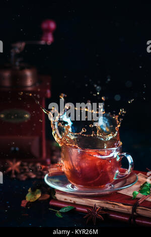 Schizzi di caffè in un bicchiere tazza da tè con il macinino da caffè, spezie, dadi e i chicchi di caffè su uno sfondo scuro. Azione fotografia bere. Foto Stock