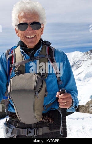 Senior uomo trekking in montagna. Viaggi avventura nelle Alpi Svizzere. Indossando occhiali da sole, camera bag bastoncini da trekking. Foto Stock