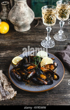 Cuocere le cozze con gusci servito sulla piastra con due bicchieri di vino bianco sul tavolo di legno Foto Stock