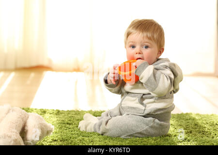 Ritratto di un bambino distratto mordere un giocattolo seduto sul pavimento a casa Foto Stock