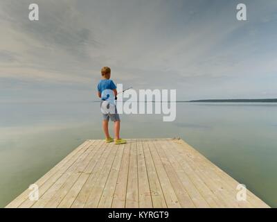 Piccoli capelli biondi del ragazzo è la pesca al fine di mole di legno. Regolare il livello di acqua nella baia, cielo chiaro. Capretto in maglietta blu, grigio striped shorts e verde fl Foto Stock