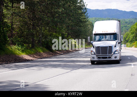 Bianco moderno big rig semi carrello con griglia cromata e dry van rimorchio il pilotaggio tramite ampia multi-linea autostrada con alberi verdi e ombra Foto Stock