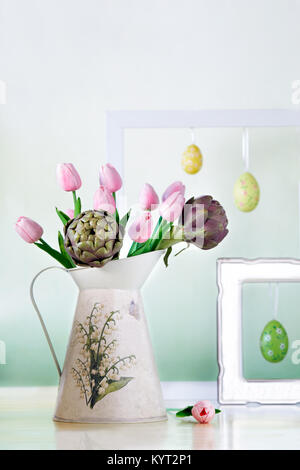 Brocca con rosa tulipani e carciofi con cornici bianche e uova di Pasqua in background. Ancora vita con molla umore in colori pastello. Foto Stock