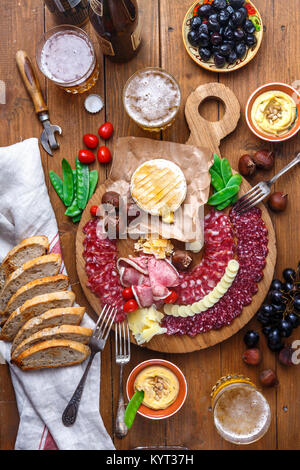 Mix di diversi spuntini e stuzzichini: salsicce, pane, olive, formaggi, castagne, piselli e birra sulla tavola di legno. Vista superiore Foto Stock