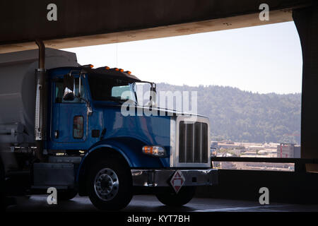 Un moderno e semplice potente blu brillante big rig semi carrello con cabina giorno intesi per il trasporto locale di merci commerciale eroga il carburante in un serbatoio Foto Stock