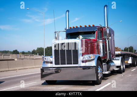 Classica big rig semi carrello in rosso scuro con alta chrome tubi e accessori per il trasporto a letto piatto semi rimorchio andando a magazzino per il carico di merci Foto Stock