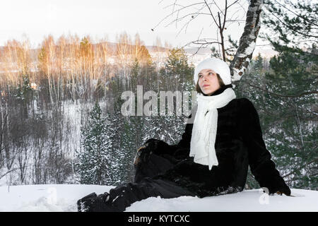 Sognante giovane ragazza si posa sulla neve in inverno soleggiata foresta di sera ed è guardando verso l'alto. Foto Stock