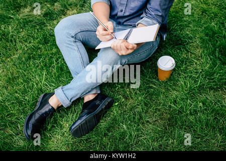 Ritagliato shot dell uomo la scrittura sul notebook e di bere il caffè dal bicchiere di carta mentre è seduto sul prato verde Foto Stock