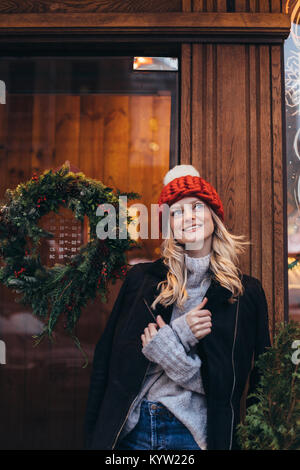 Giovane donna bionda in rosso berretto lavorato a maglia con pompon bianco, grigio maglione, giacca nera e jeans blu fare in posa con Christmac cafe finestra sul backg Foto Stock