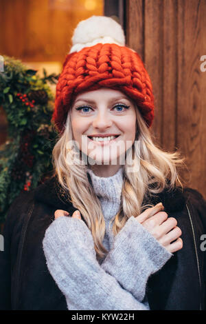 Giovani dagli occhi blu donna bionda in rosso berretto lavorato a maglia con pompon bianco, grigio maglione e giacca nera fare in posa con Christmac cafe finestra sul backgro Foto Stock