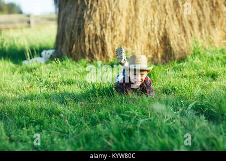 Bello piccolo ragazzo giace sull'erba su sfondo di pagliaio. Bambino riposa in natura, giace sul suo stomaco e propps la testa con le mani. Ritratto di giovane Foto Stock