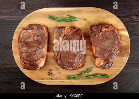 Una foto aerea di tre fette di carne cotta, filetti di manzo, ripresa dall'alto sul buio rustica con rametti di rosmarino, sale e pepe Foto Stock