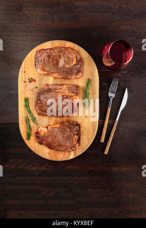 Una foto aerea di tre fette di carne cotta, filetti di manzo, ripresa dall'alto sul buio rustica con rametti di rosmarino, sale, pepe, vino, Foto Stock