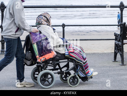 Persona più giovane (care/famiglia?) spingendo con sedia a rotelle donna anziana lungo il lungomare. Regno Unito Foto Stock