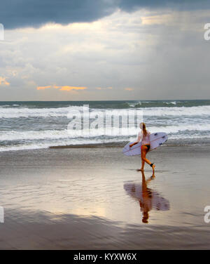 Donna surfer di camminare sulla spiaggia con la tavola da surf al tramonto. Isola di Bali