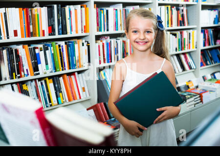 Ragazza bionda in età scolare tenendo il libro prescelto in mani in bookstore Foto Stock