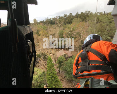 Un membro di equipaggio guarda sulle case e strade di un quartiere colpiti dalla Santa Barbara County colate di fango a Santa Barbara in California sono mostrati, gen. 9, 2018, dal punto di vista di un Coast Guard MH-65 Delfino elicottero equipaggio coinvolto nel salvataggio di feriti e vittime a filamento. Coast Guard elicotteri sono stati spediti da Los Angeles-Long Beach e San Diego per assistere i primi responder con gli sforzi di salvataggio. (U.S. Coast Guard Foto Stock