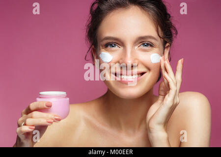 Donna l'applicazione di crema antirughe o anti-invecchiamento della pelle crema di cura. Foto di donna sorridente con pelle sana su sfondo rosa. Cura della Pelle concept Foto Stock