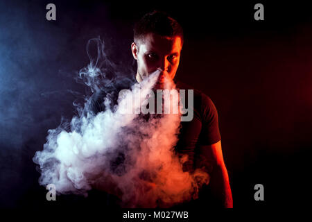 Giovane maschio elegante smoker in black T-shirt tirate fuori il suo naso e una grande nuvola di fumo da vape su un nero sfondo isolato Foto Stock