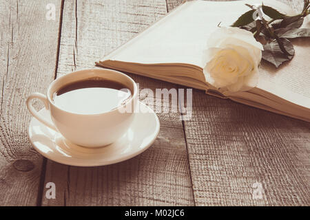 White tazza di caffè nero e libro aperto con rosa bianca sulla tavola di legno. Vintage tonica. Close up. Foto Stock