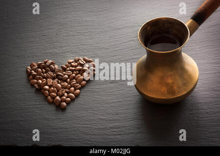 Nero caffè in rame antico cezve e i chicchi di caffè in forma di cuore su nero ardesia come sfondo. Caffè vecchio pentola su sfondo scuro. Foto Stock
